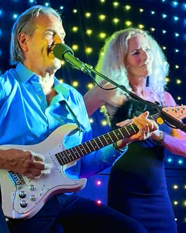 Ron Rox singing and playing guitar with Toni René beside him, in front of a curtain of stage lights